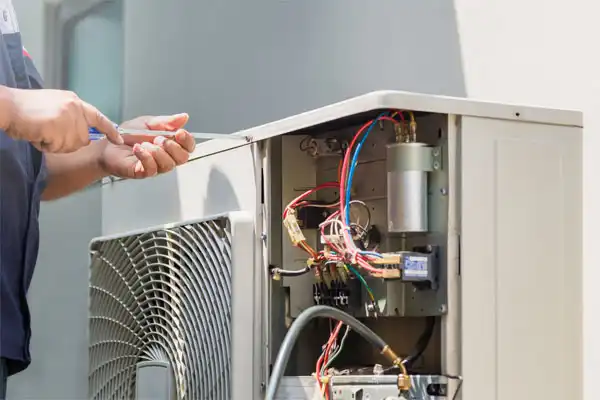 Technician repairing an outdoor air conditioning unit, with wires exposed and tools in hand.
