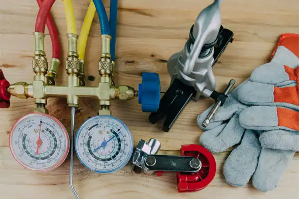 HVAC tools on a wooden surface: pressure gauges with hoses, bending tool, cutter, wrench, and a pair of work gloves.