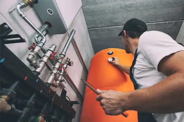 A worker in a cap and apron inspects an orange tank in a utility room, surrounded by pipes and valves.