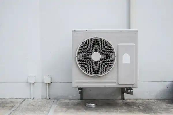 Outdoor air conditioning unit mounted on a wall with visible pipes and wires on the left.