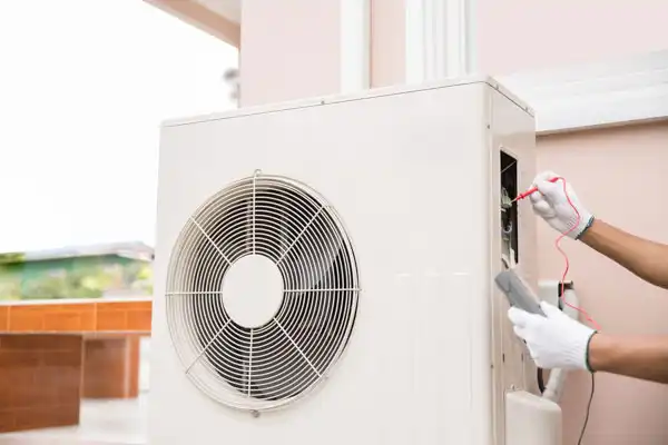 A person in gloves uses a multimeter to test an outdoor air conditioning unit.
