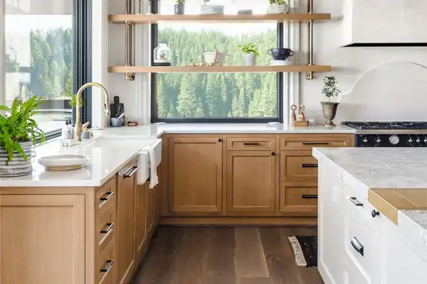 A modern kitchen with wooden cabinets, white countertops, and a view of a forest through the window. There are plants on the shelves, a gold faucet, and a black oven.