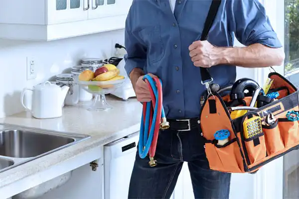 A person in a blue shirt holds a tool bag and colorful hoses in a kitchen next to a sink and fruit bowl.