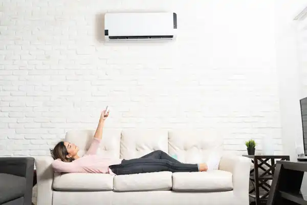 Person lying on a beige sofa, pointing a remote at an air conditioner mounted on a white brick wall.