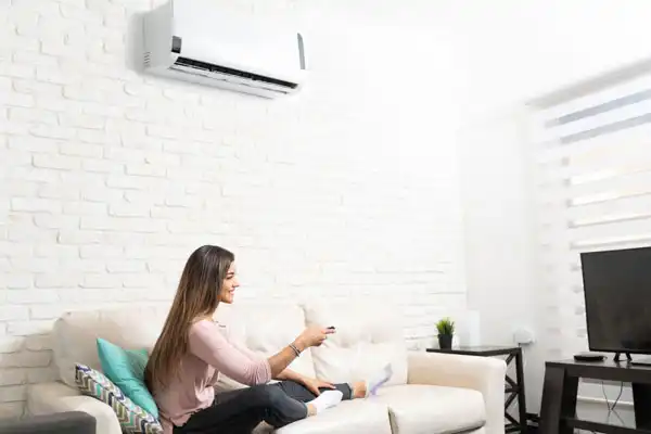 A woman sits on a sofa in a bright living room, using a remote control. An air conditioner is mounted on the white brick wall above her. A TV is placed on a stand nearby.