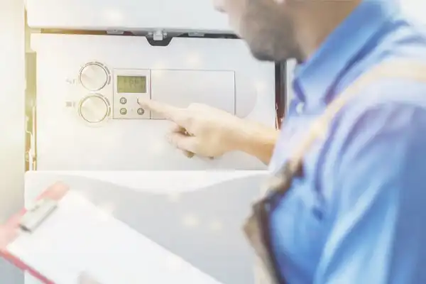 A person in a blue shirt adjusts the settings on a home boiler control panel, holding a clipboard in the other hand.