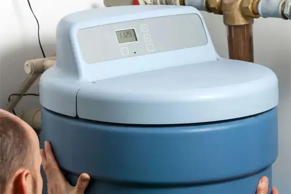 Person adjusting a blue water softener unit with a digital display and buttons, connected to pipes.