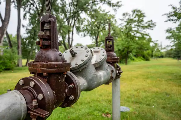 A metal valve assembly is mounted on a pipe in a grassy field. Trees are visible in the background under an overcast sky.
