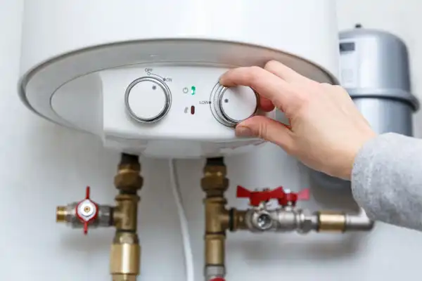 Hand adjusting the temperature dial on a water heater with visible pipes and valves.