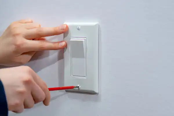 Hands using a screwdriver to adjust or install a white light switch on a wall.